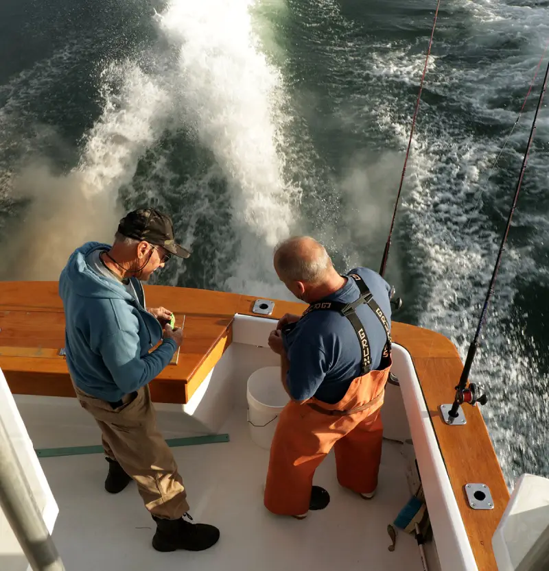 os3 Two fishermen on a boat, preparing gear with a scenic view of waves splashing behind them under warm sunlight.