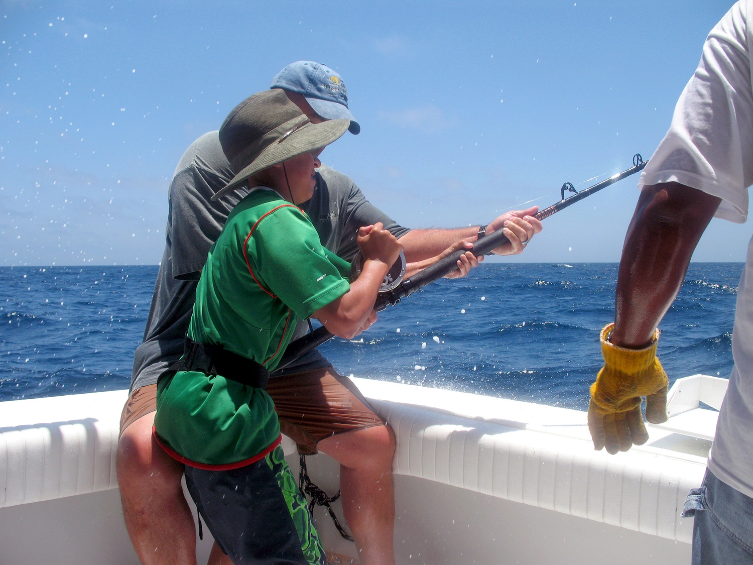 A child in a green shirt holding a fishing rod, assisted by an adult, on a boat surrounded by splashing water and blue ocean A child in a green shirt holding a fishing rod, assisted by an adult, on a boat surrounded by splashing water and blue ocean